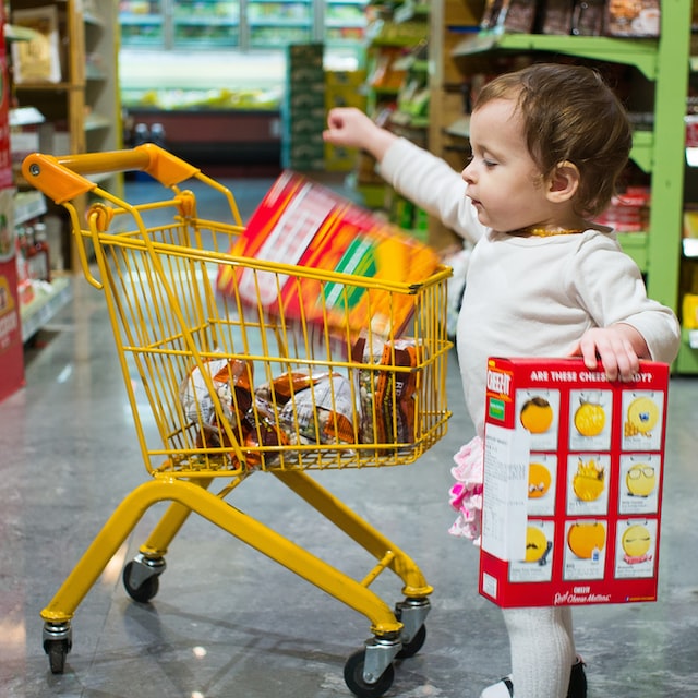 Ein Kleinkind steht im Supermarkt seitlich vor einem kleinen gelben Kinder-Einkaufswagen und wirft eine große Schachtel hinein, eine zweite hält es in der anderen Hand.