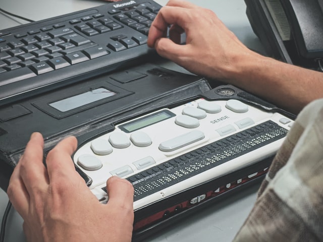 Zwei Hände bedienen eine schwarze Computertastatur und eine Braille-Maschine, die darunter liegt.